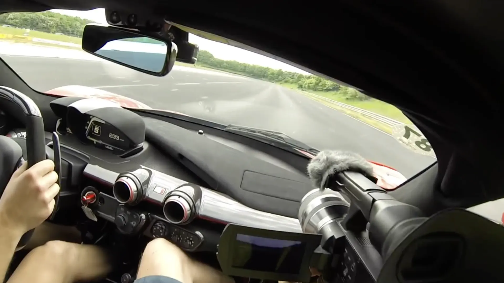 View from the passenger seat of a Ferrari LaFerrari Aperta on a straight. The display reads sixth gear and 233 km/h. One of the driver's hands rests on the wheel.
